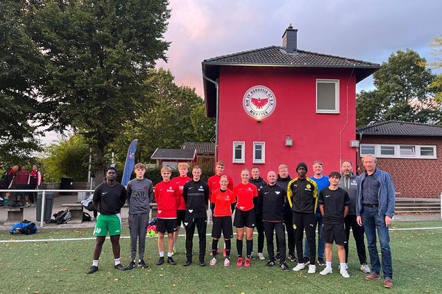 Gruppe von Teenagern in Sportkleidung steht vor einem Vereinsgeb&auml;ude auf einem Fu&szlig;ballplatz.