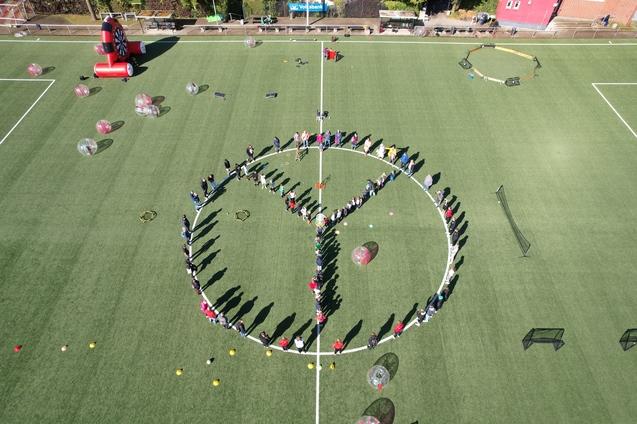 Menschen bilden einen gro&szlig;en Kreis mit einem Peace-Zeichen auf einem Fu&szlig;ballfeld, umgeben von bunten Ballons und Equipment.