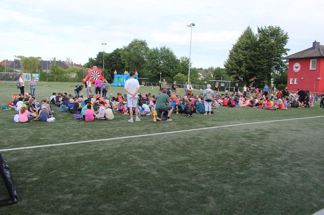 Viele Kinder sitzen auf einem Sportplatz, w&auml;hrend eine Veranstaltung mit bunten Attraktionen im Hintergrund stattfindet.