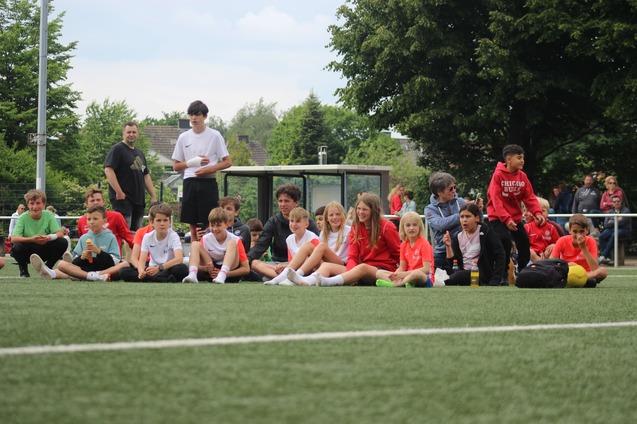 Gruppe von Kindern und Erwachsenen sitzend am Rande eines Fu&szlig;ballfeldes, umgeben von gr&uuml;ner Natur.
