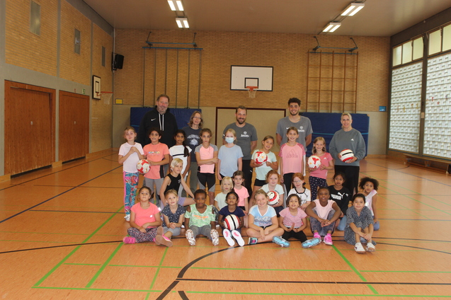 Gruppenfoto von Kindern und Trainern in einer Sporthalle, einige halten Volleyballs, umgeben von Holzfu&szlig;boden und W&auml;nden.