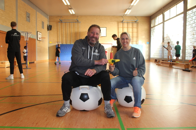 Zwei Personen sitzen auf gro&szlig;en, runden Fu&szlig;b&auml;llen in einer Sporthalle und l&auml;cheln in die Kamera.