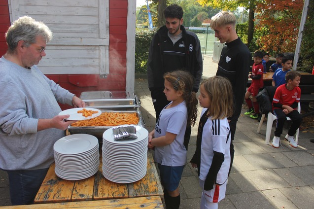 Ein Mann serviert Pasta an Kinder in Sportkleidung vor einem Tisch mit wei&szlig;en Tellern im Freien.