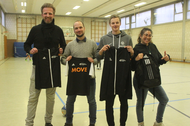 Vier Personen in Sportkleidung halten T-Shirts mit dem Schriftzug &bdquo;MOVE&ldquo; in einer Sporthalle.