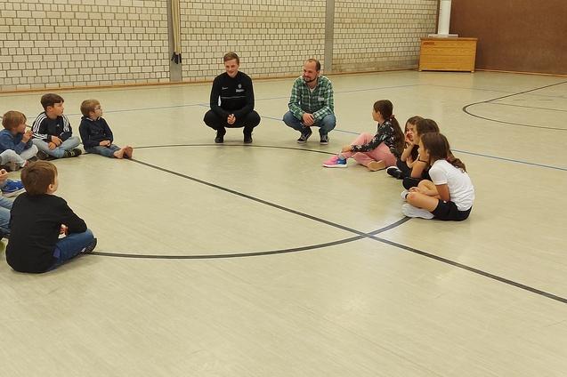 Zwei Erwachsene sitzen auf dem Boden, umgeben von einer Gruppe von Kindern in einer Sporthalle.