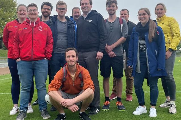 Gruppe von zehn Personen l&auml;chelt auf einem gr&uuml;nen Rasenplatz, einige tragen Sportjacken und stehen in lockerer Pose.