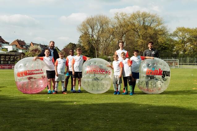 Gruppe von Kindern und Erwachsenen in Zorb-B&auml;llen auf einem Fu&szlig;ballfeld, umgeben von B&auml;umen und einem Sportplatz.