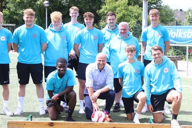 Gruppenfoto von zehn Fu&szlig;ballspielern in blauen Shirts und zwei Betreuern auf einem Sportplatz mit Banner im Hintergrund.