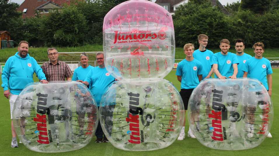 Gruppenszene von Personen in blauen T-Shirts neben gro&szlig;en, transparenten Bubble-Soccer-B&auml;llen auf einem Rasenplatz.