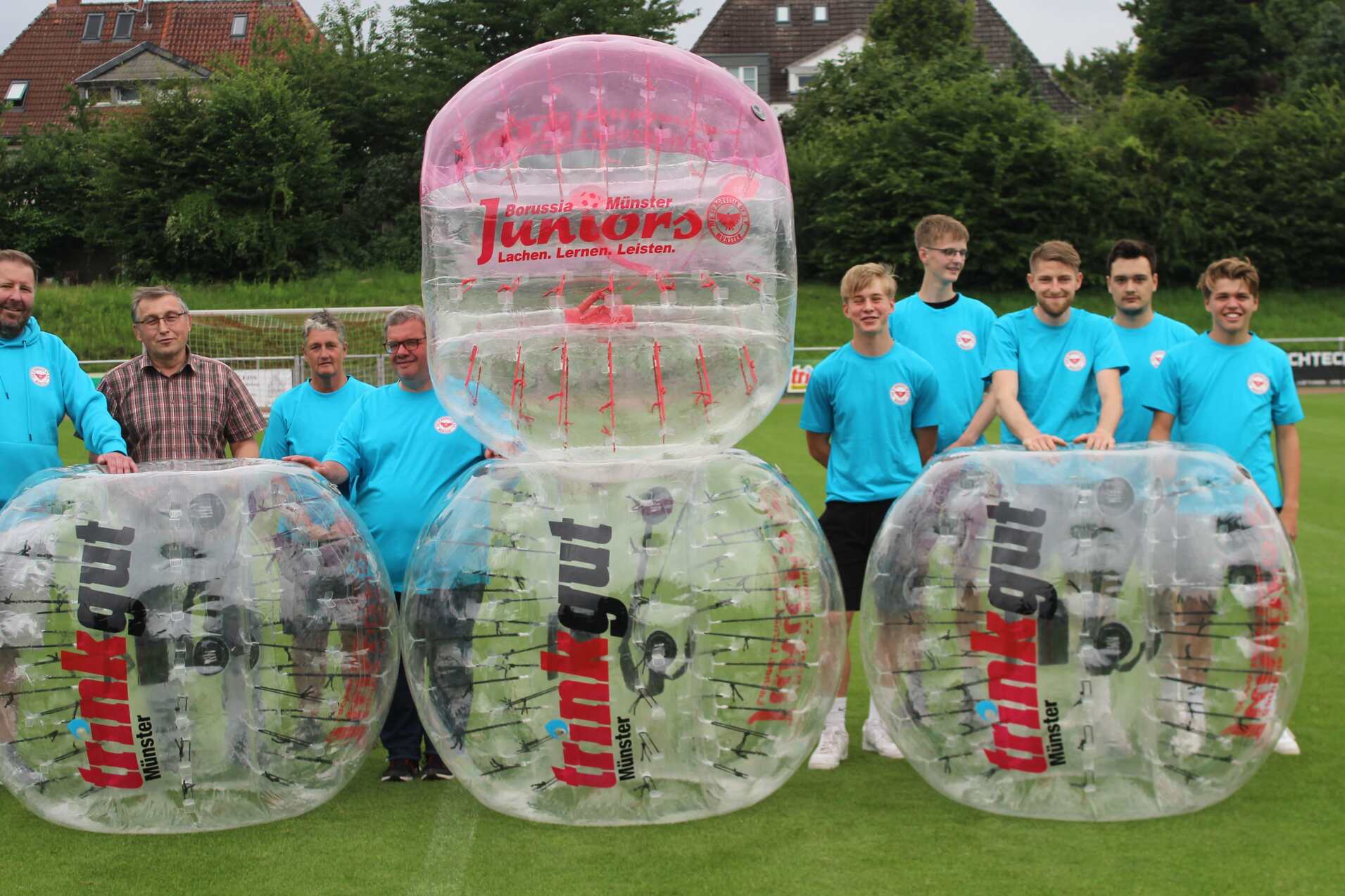 Gruppenszene von Personen in blauen T-Shirts neben gro&szlig;en, transparenten Bubble-Soccer-B&auml;llen auf einem Rasenplatz.