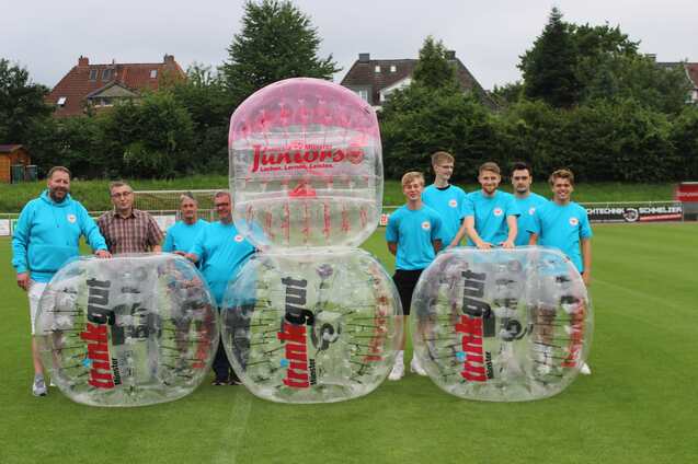 Gruppenszene von Personen in blauen T-Shirts neben gro&szlig;en, transparenten Bubble-Soccer-B&auml;llen auf einem Rasenplatz.