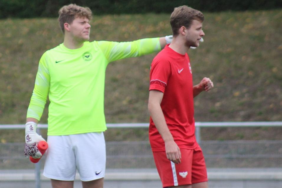 Zwei Fu&szlig;ballspieler stehen auf dem Platz; der Torwart in leuchtend gr&uuml;nem Trikot legt die Hand auf die Schulter eines Mitspielers.