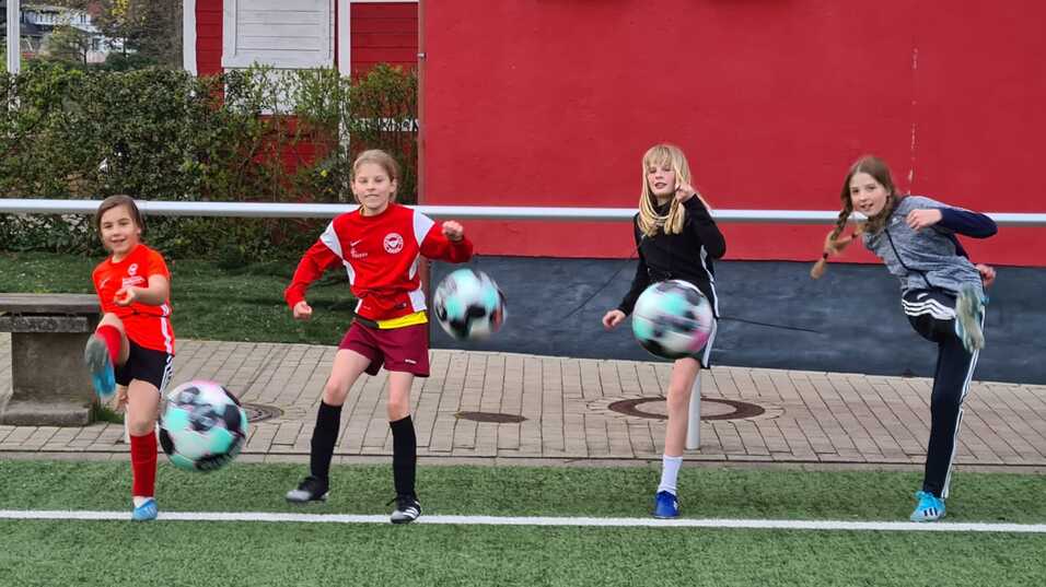 Vier Kinder stehen auf einem Fu&szlig;ballplatz und halten bunte Fu&szlig;b&auml;lle, hinter ihnen ist eine rote Wand zu sehen.