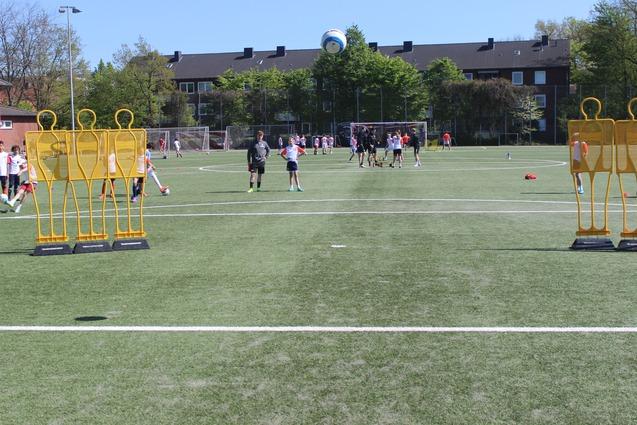 Kinder trainieren auf einem Fu&szlig;ballplatz, umgeben von Trainingspuppen und einer blauen Ball, im Hintergrund Wohngeb&auml;ude.