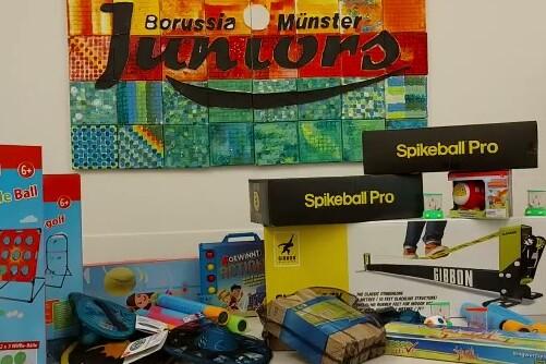 Bunte Spielzeugartikel und Sportger&auml;te auf einem Tisch, mit dem Banner &bdquo;Borussia M&uuml;nster Juniors&ldquo; im Hintergrund.