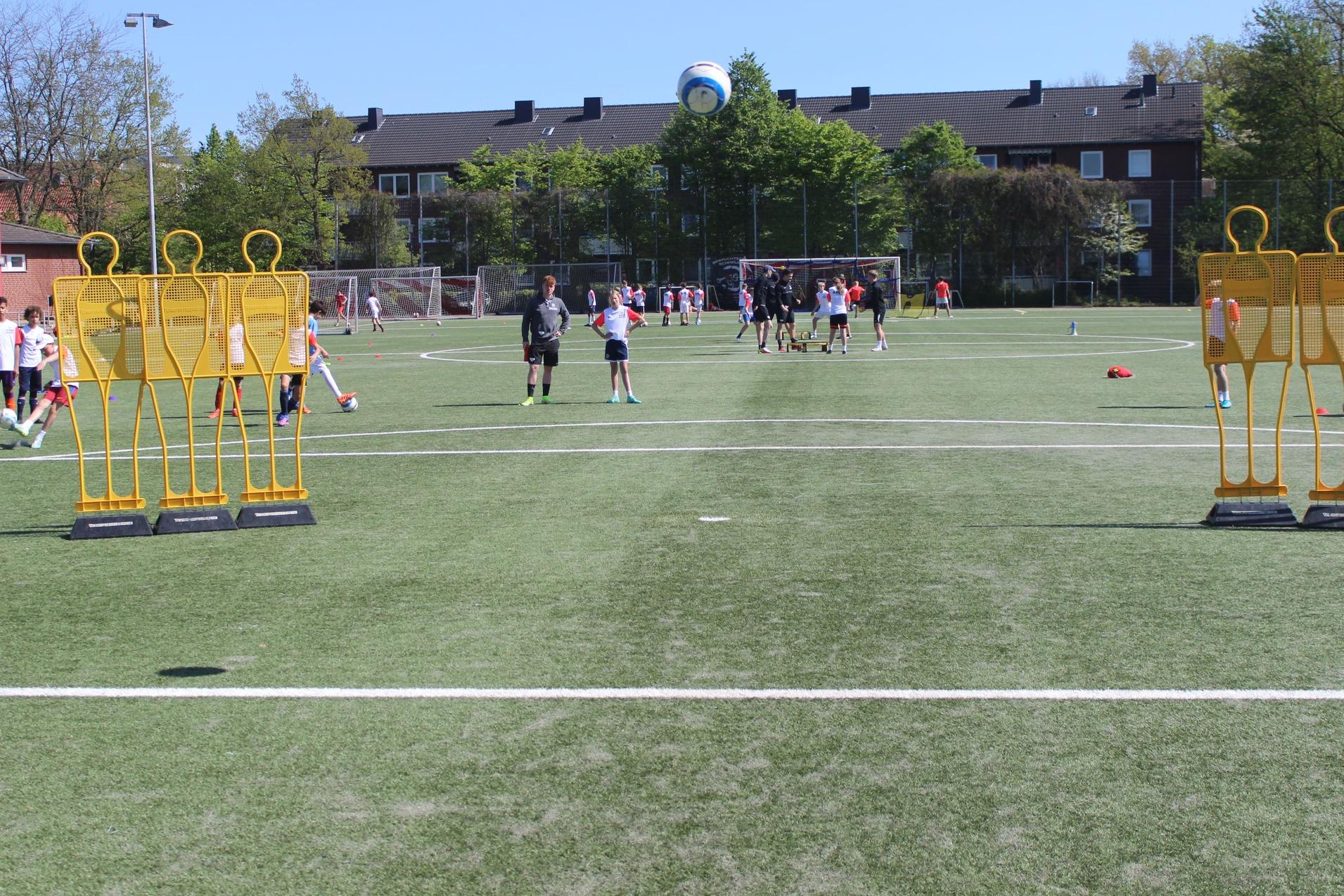 Kinder trainieren auf einem Fu&szlig;ballplatz, umgeben von Trainingspuppen und einer blauen Ball, im Hintergrund Wohngeb&auml;ude.