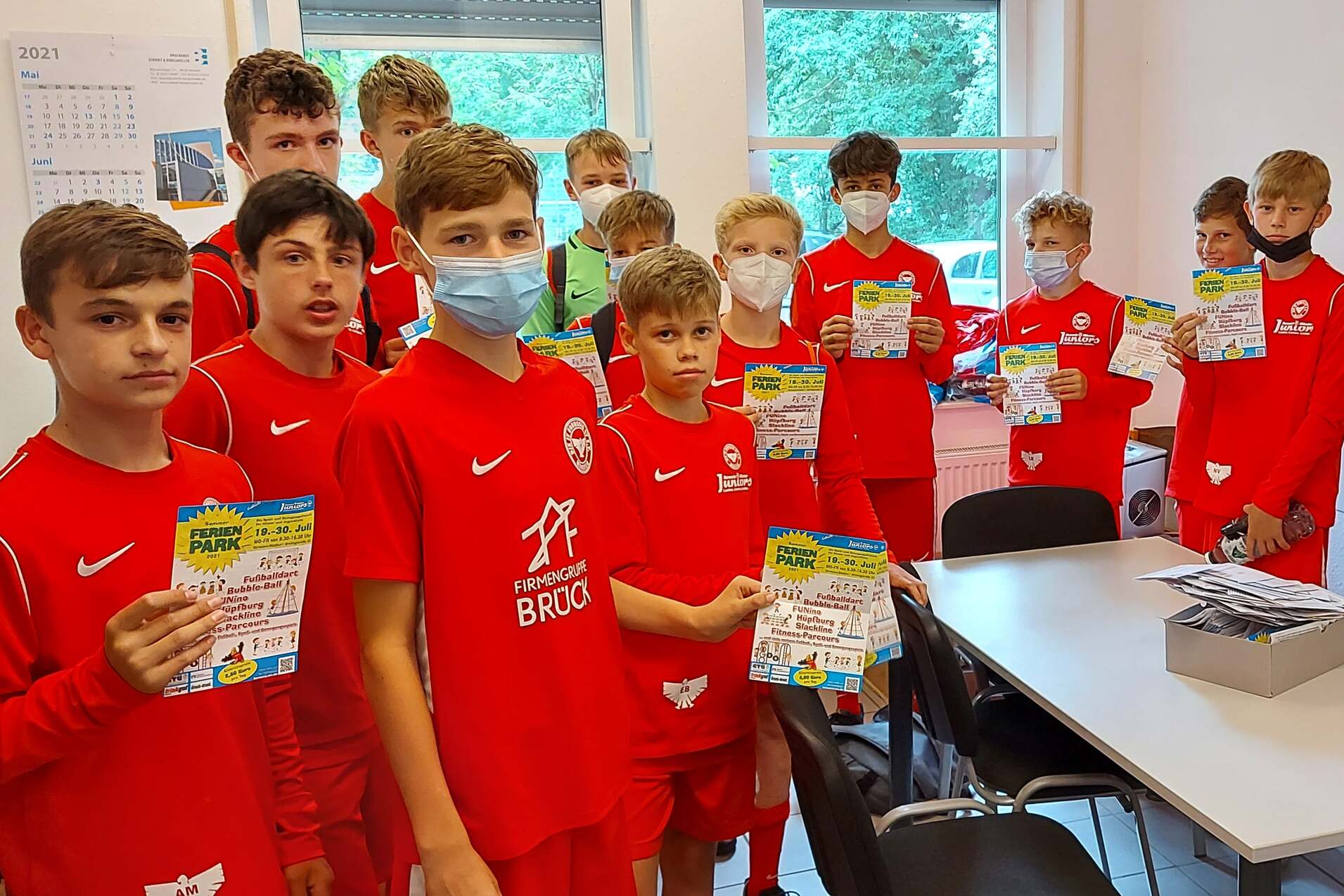Eine Gruppe von Jungen in roten Sportanz&uuml;gen h&auml;lt Dokumente in einem Raum mit Tischen und Fenstern.