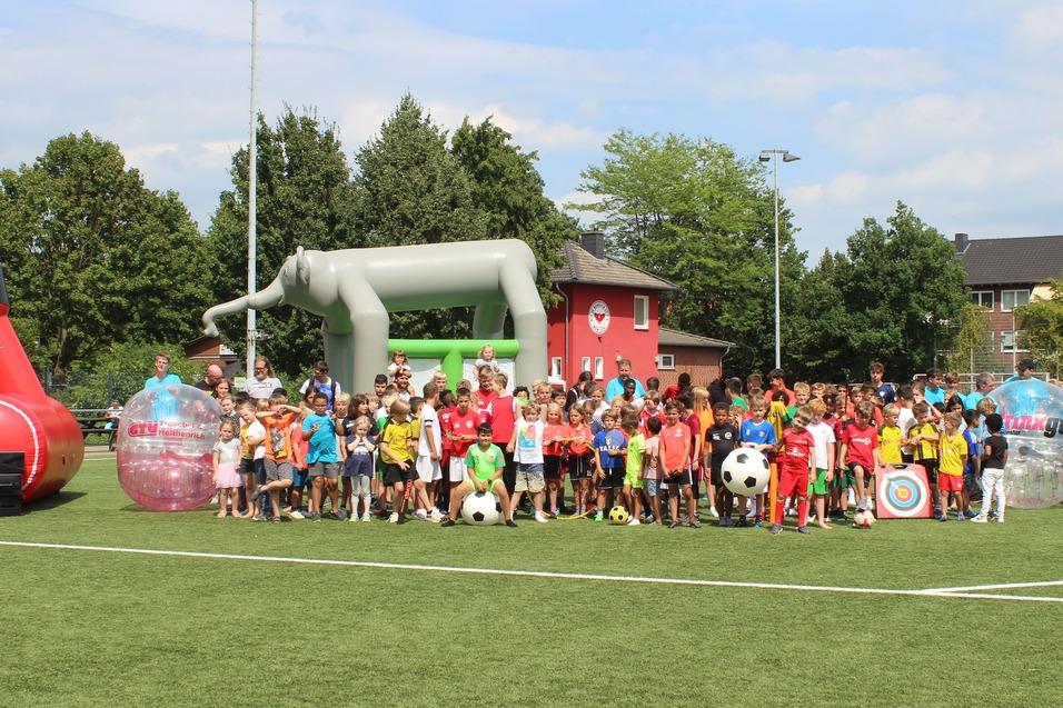 Viele Kinder stehen auf einem Fu&szlig;ballplatz, umgeben von Spielger&auml;ten und einem bunten Hintergrund.