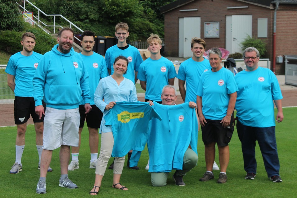 Gruppe von zehn Personen in blauen T-Shirts auf einem Sportplatz, lachend und posierend f&uuml;r das Foto.