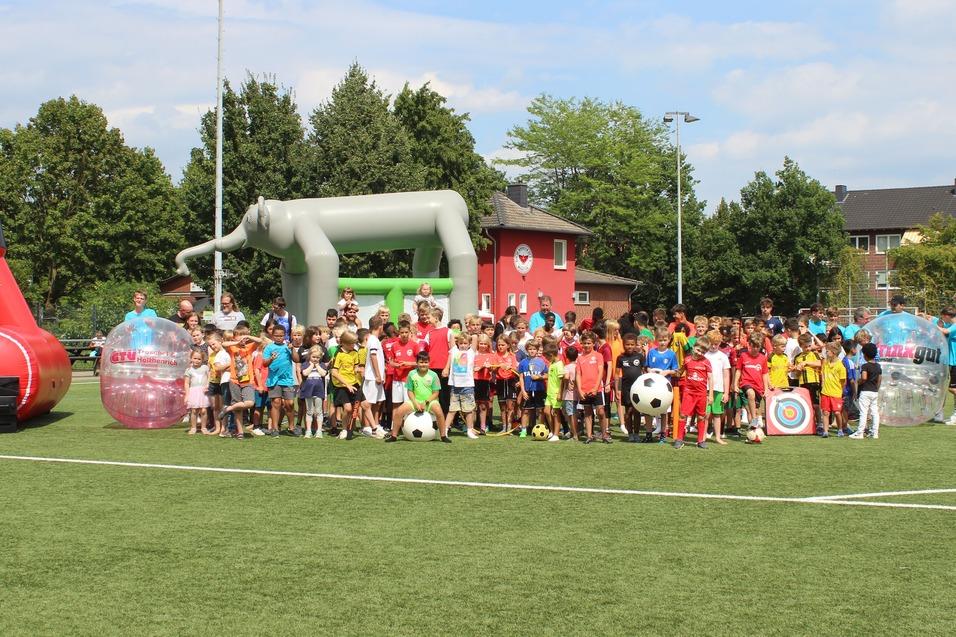 Viele Kinder stehen auf einem Fu&szlig;ballplatz, umgeben von Spielger&auml;ten und einem bunten Hintergrund.