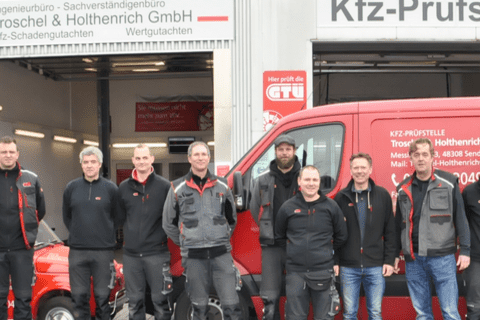 Gruppe von Mitarbeitenden vor einer KFZ-Pr&uuml;fstelle, mit einem roten Lieferwagen und einem Oldtimer im Hintergrund.
