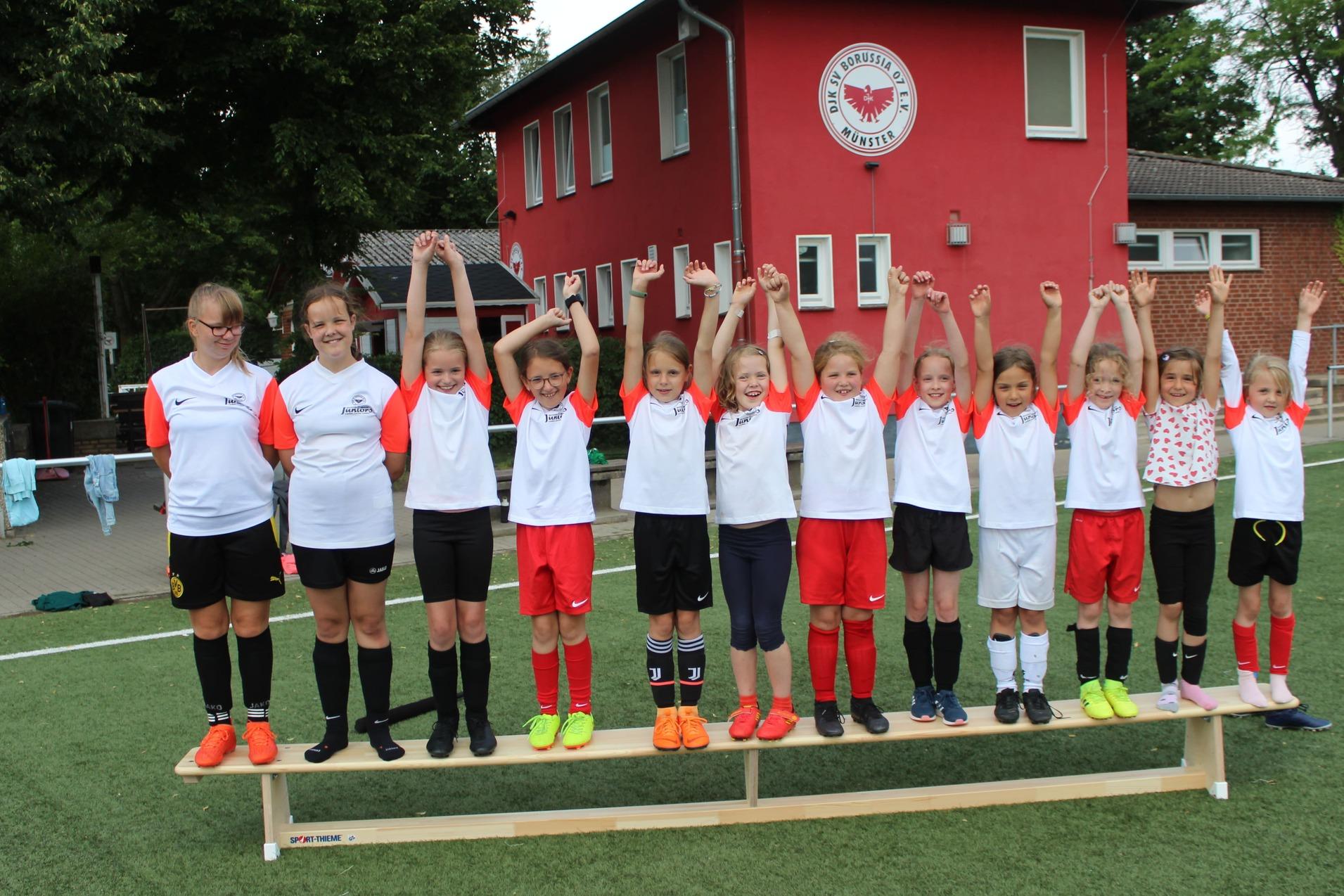 Gruppenfoto von 12 M&auml;dchen in Sporttrikots, die auf einer Bank stehen und ihre Arme jubelnd in die Luft heben.