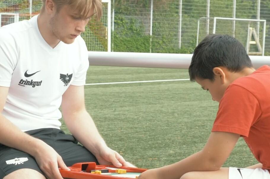 Zwei Jungen sitzen auf einer Bank und spielen ein Brettspiel auf einem Fu&szlig;ballplatz im Freien.