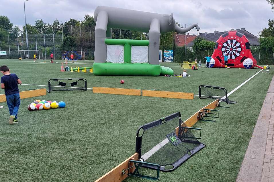Auf einem Sportplatz stehen ein gr&uuml;nes H&uuml;pfburg und ein Ziel-Dartspiel, w&auml;hrend Kinder spielen und Erwachsene sitzen.
