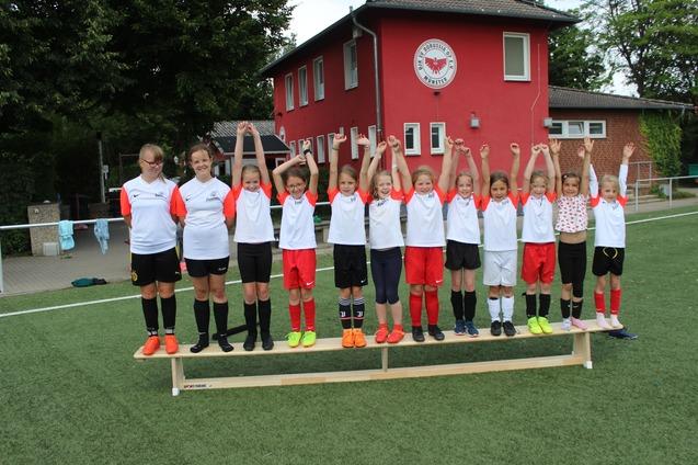 Gruppenfoto von 12 M&auml;dchen in Sporttrikots, die auf einer Bank stehen und ihre Arme jubelnd in die Luft heben.