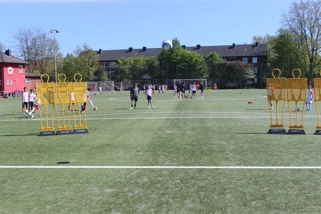 Kinder trainieren auf einem Fu&szlig;ballplatz, umgeben von Trainingspuppen und einer blauen Ball, im Hintergrund Wohngeb&auml;ude.