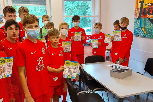 Eine Gruppe von Jungen in roten Sportanz&uuml;gen h&auml;lt Dokumente in einem Raum mit Tischen und Fenstern.