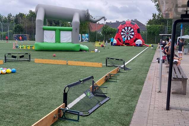 Auf einem Sportplatz stehen ein gr&uuml;nes H&uuml;pfburg und ein Ziel-Dartspiel, w&auml;hrend Kinder spielen und Erwachsene sitzen.