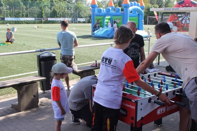 Kinder und Erwachsene spielen an einem Tischkickertisch auf einem Sportplatz mit Spielger&auml;ten im Hintergrund.