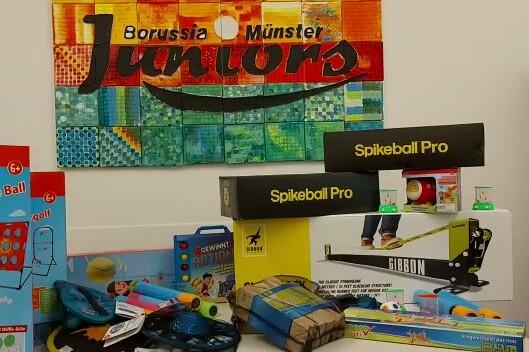 Bunte Spielzeugartikel und Sportger&auml;te auf einem Tisch, mit dem Banner &bdquo;Borussia M&uuml;nster Juniors&ldquo; im Hintergrund.