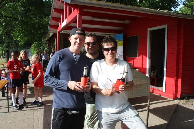 Drei Personen stehen vor einem roten Kiosk und halten Flaschen in den H&auml;nden, im Hintergrund sind Kinder in Sportbekleidung zu sehen.