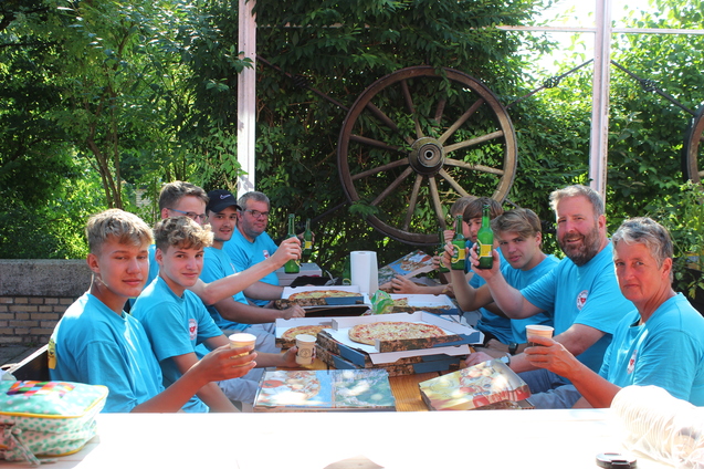 Gruppe von acht Personen in blauen T-Shirts feiert mit Pizza und Getr&auml;nken an einem Tisch im Freien.