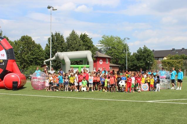 Viele Kinder stehen auf einem Fu&szlig;ballplatz, umgeben von Spielger&auml;ten und einem bunten Hintergrund.