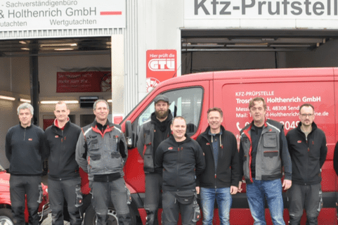 Gruppe von Mitarbeitenden vor einer KFZ-Pr&uuml;fstelle, mit einem roten Lieferwagen und einem Oldtimer im Hintergrund.