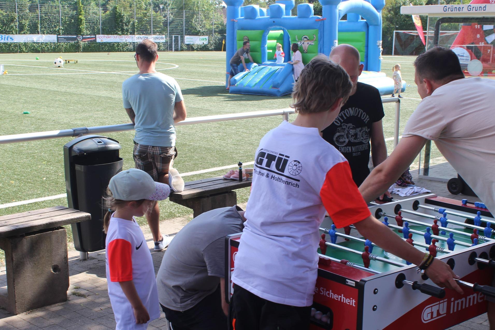 Kinder und Erwachsene spielen an einem Tischkickertisch auf einem Sportplatz mit Spielger&auml;ten im Hintergrund.