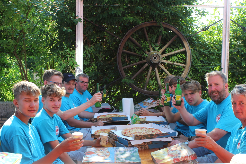 Gruppe von acht Personen in blauen T-Shirts feiert mit Pizza und Getr&auml;nken an einem Tisch im Freien.