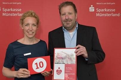 Zwei Personen halten ein Zertifikat und eine Nummer 10 vor einem roten Hintergrund mit dem Logo der Sparkasse.