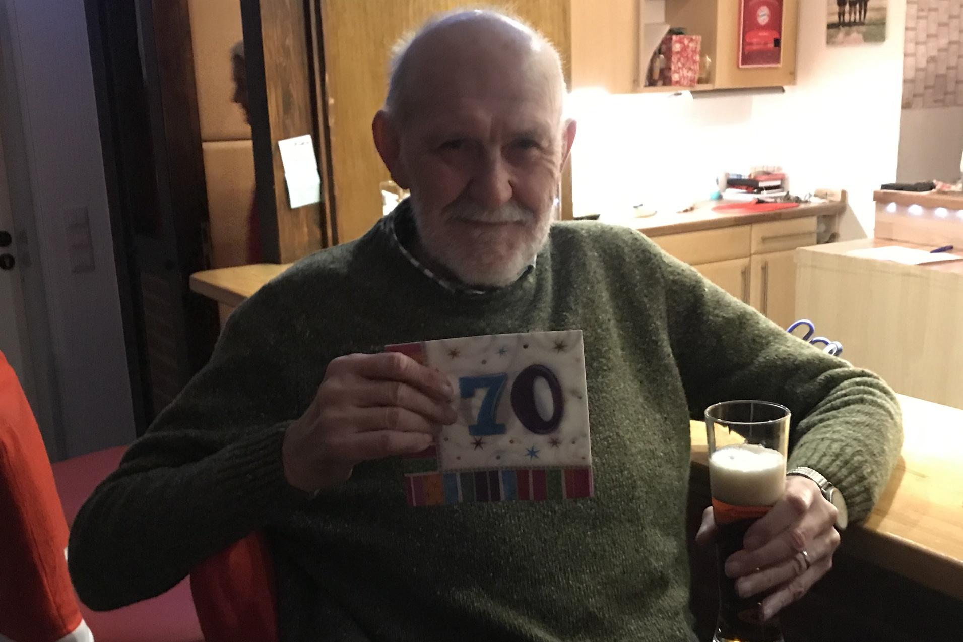 Älterer Mann in einem grünen Pullover hält eine „70“-Glückwunschkarte und ein Bierglas in einer gemütlichen Umgebung.