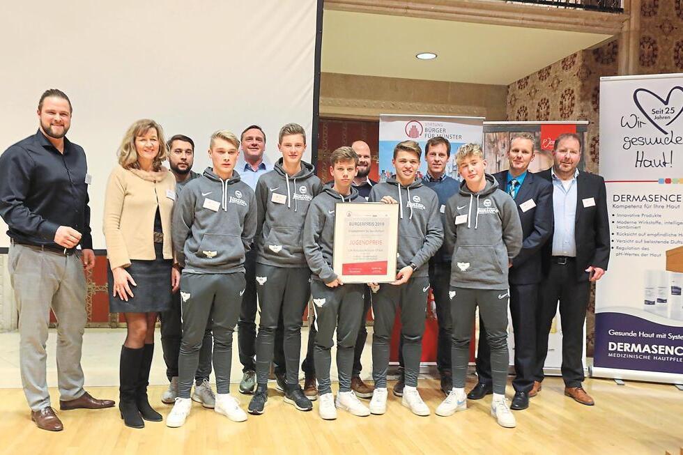 Gruppe von Personen in Sportkleidung steht auf einer B&uuml;hne, w&auml;hrend ein Zertifikat &uuml;berreicht wird.