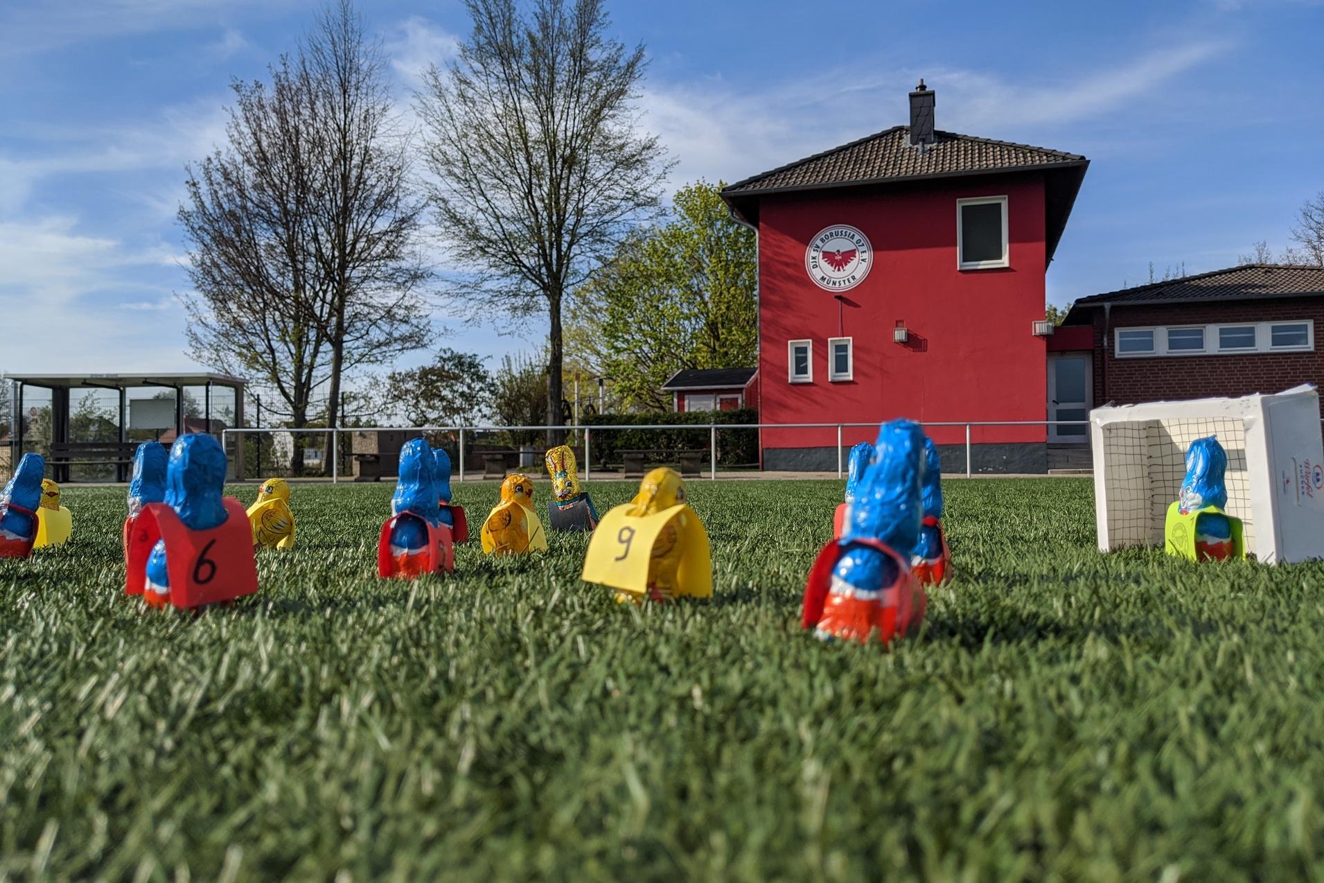 Bunte Spielfiguren in Form von Spielern stehen auf einem Fussballfeld vor einem roten Vereinsgeb&auml;ude.