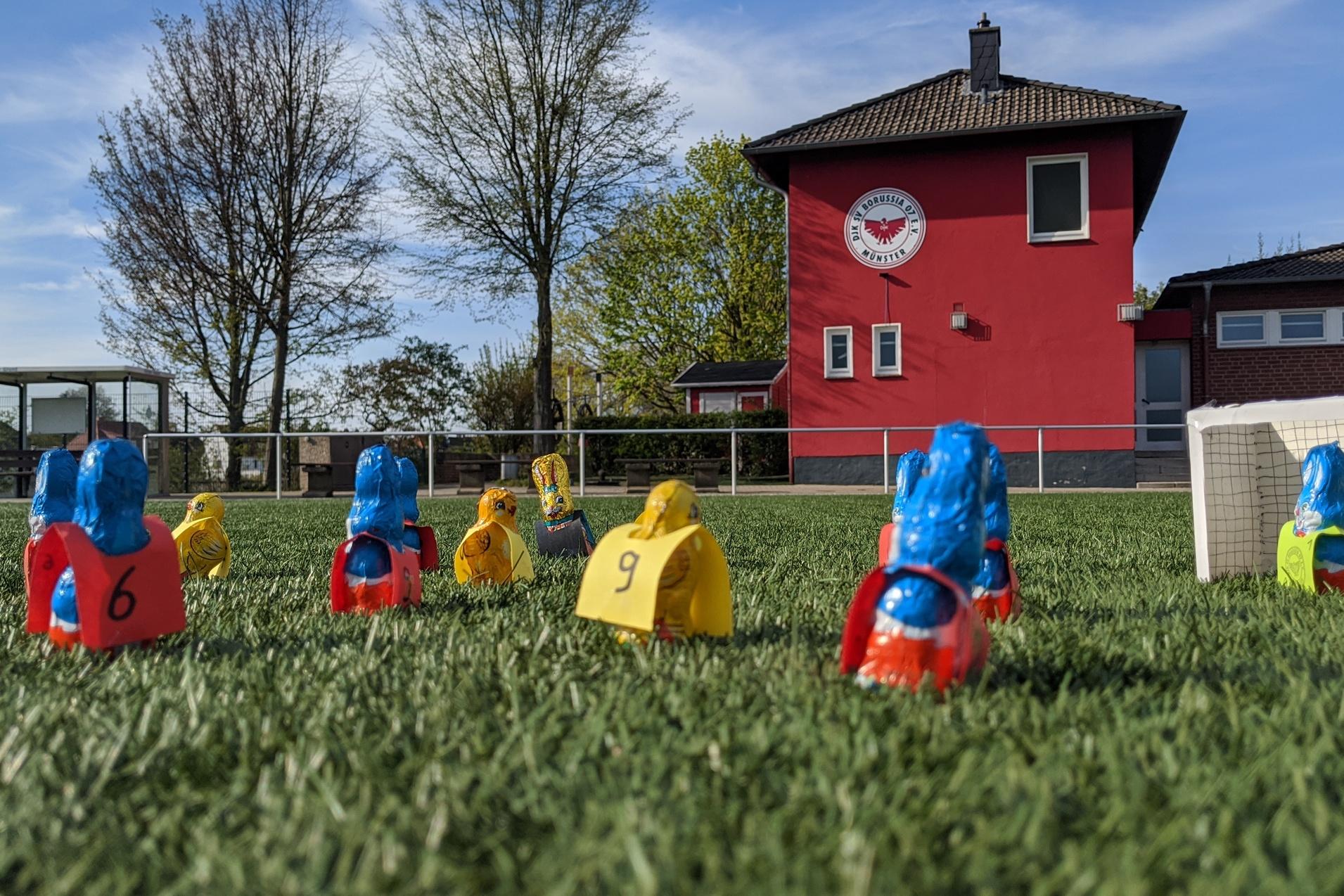 Bunte Spielfiguren in Form von Spielern stehen auf einem Fussballfeld vor einem roten Vereinsgebäude.