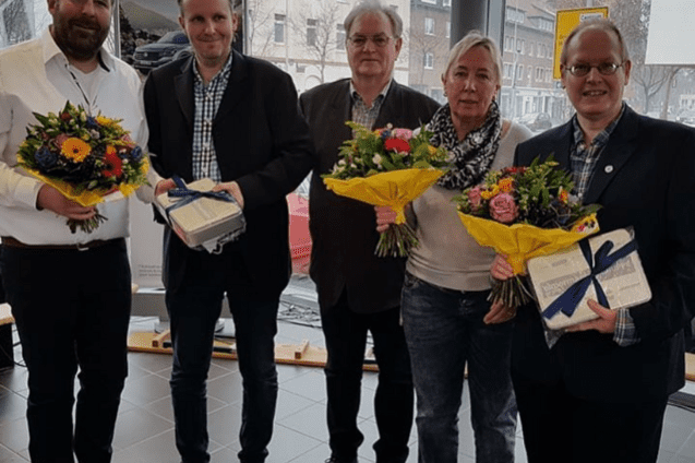 Gruppe von f&uuml;nf Personen, die Blumenstr&auml;u&szlig;e und Geschenke in einem hellen Raum halten. Fenster im Hintergrund.