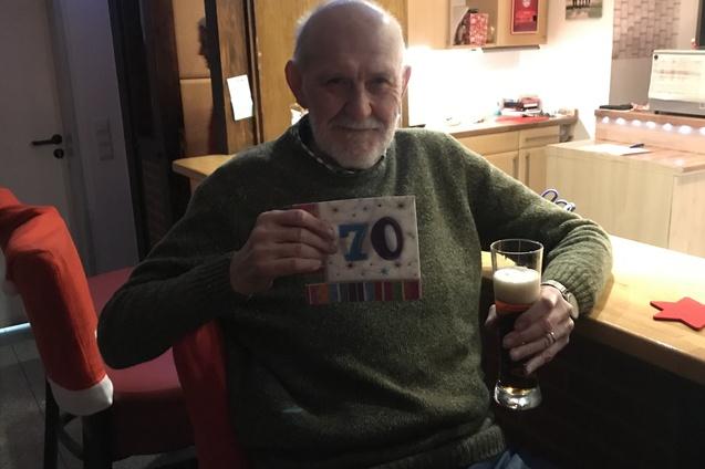 &Auml;lterer Mann in einem gr&uuml;nen Pullover h&auml;lt eine &bdquo;70&ldquo;-Gl&uuml;ckwunschkarte und ein Bierglas in einer gem&uuml;tlichen Umgebung.