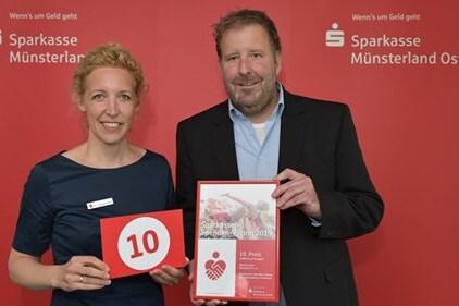 Zwei Personen halten ein Zertifikat und eine Nummer 10 vor einem roten Hintergrund mit dem Logo der Sparkasse.
