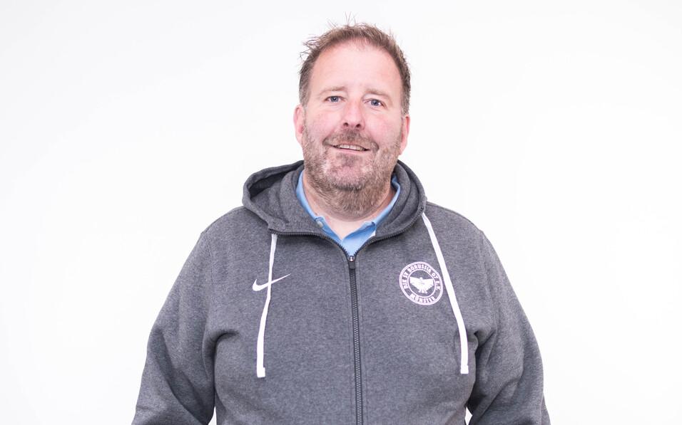 Mann mit Bart in grauem Hoodie steht vor einem wei&szlig;en Hintergrund und l&auml;chelt freundlich in die Kamera.