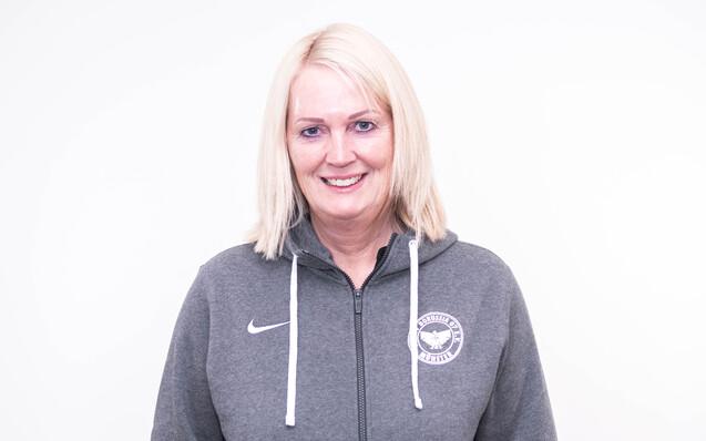 Frau mit blonden, schulterlangen Haaren in grauem Sportpullover, l&auml;chelt vor einem wei&szlig;en Hintergrund.